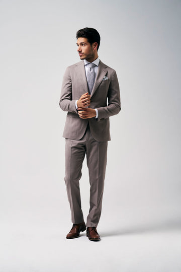 Mannelijk model in een taupegrijs kostuum met stropdas en pochet, elegant poserend tegen een lichte achtergrond.