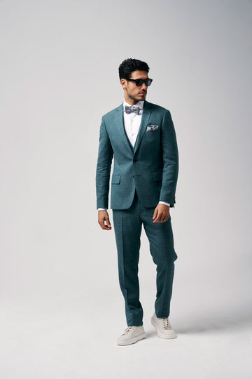 Mannelijk model in een groenblauw pak met strik en zonnebril, staand tegen een lichte achtergrond.