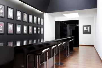 Modern en strak interieur met een lange zwarte Wellens bar, hoge barkrukken en ingelijste foto’s aan de muur, afgewerkt met een houten vloer en minimalistische verlichting.